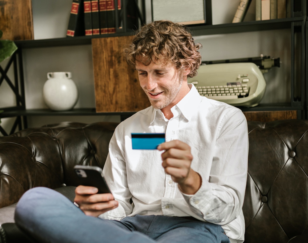 image of a white man shitting on a brown leather couch looking at his phone to enter credit card information into an ecommerce website