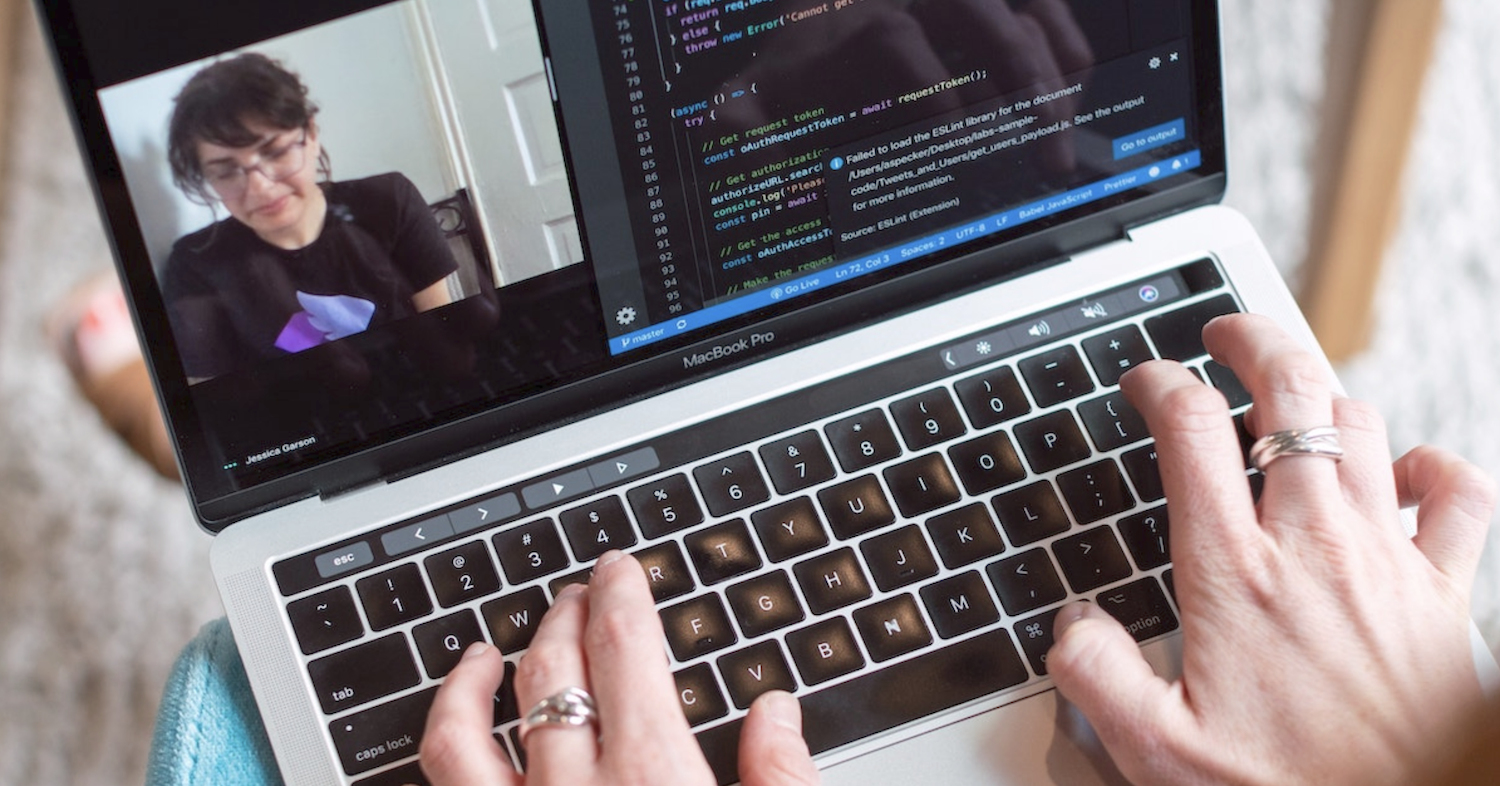 decorative image of a laptop in someones lap; you can see their hands which seem to by typing. On the screen is a video call with a woman on it, next to a code editing screen
