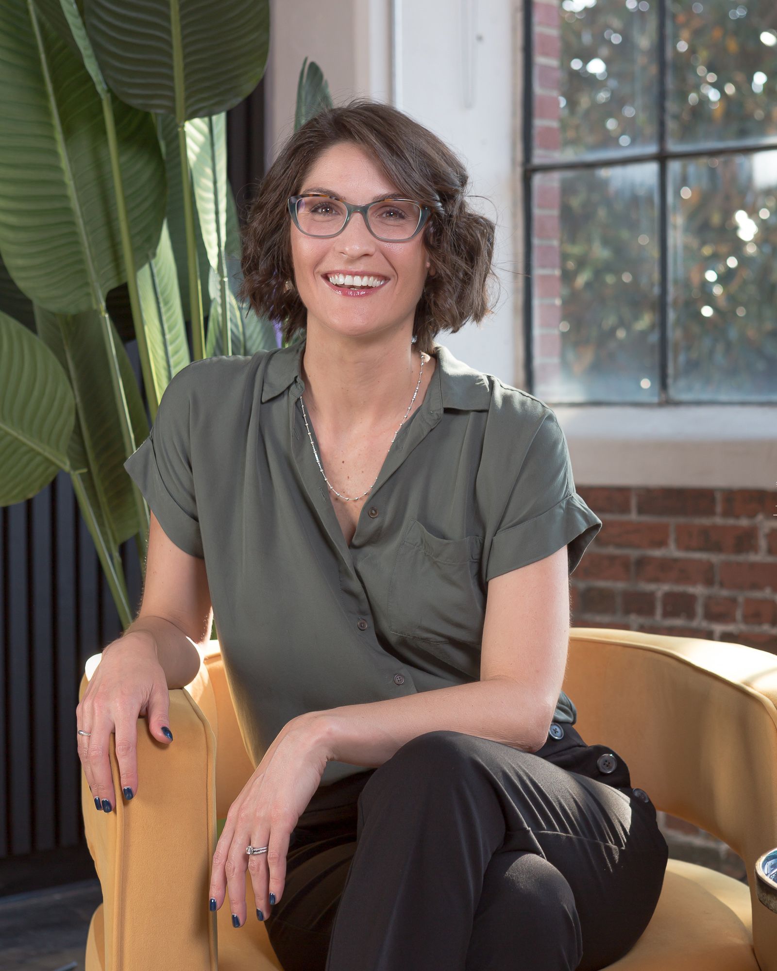 Headshot of Tory Gray with short brown hair and glasses, wearing a green shirt, sits smiling on a yellow chair with a leafy plant and window in the background.