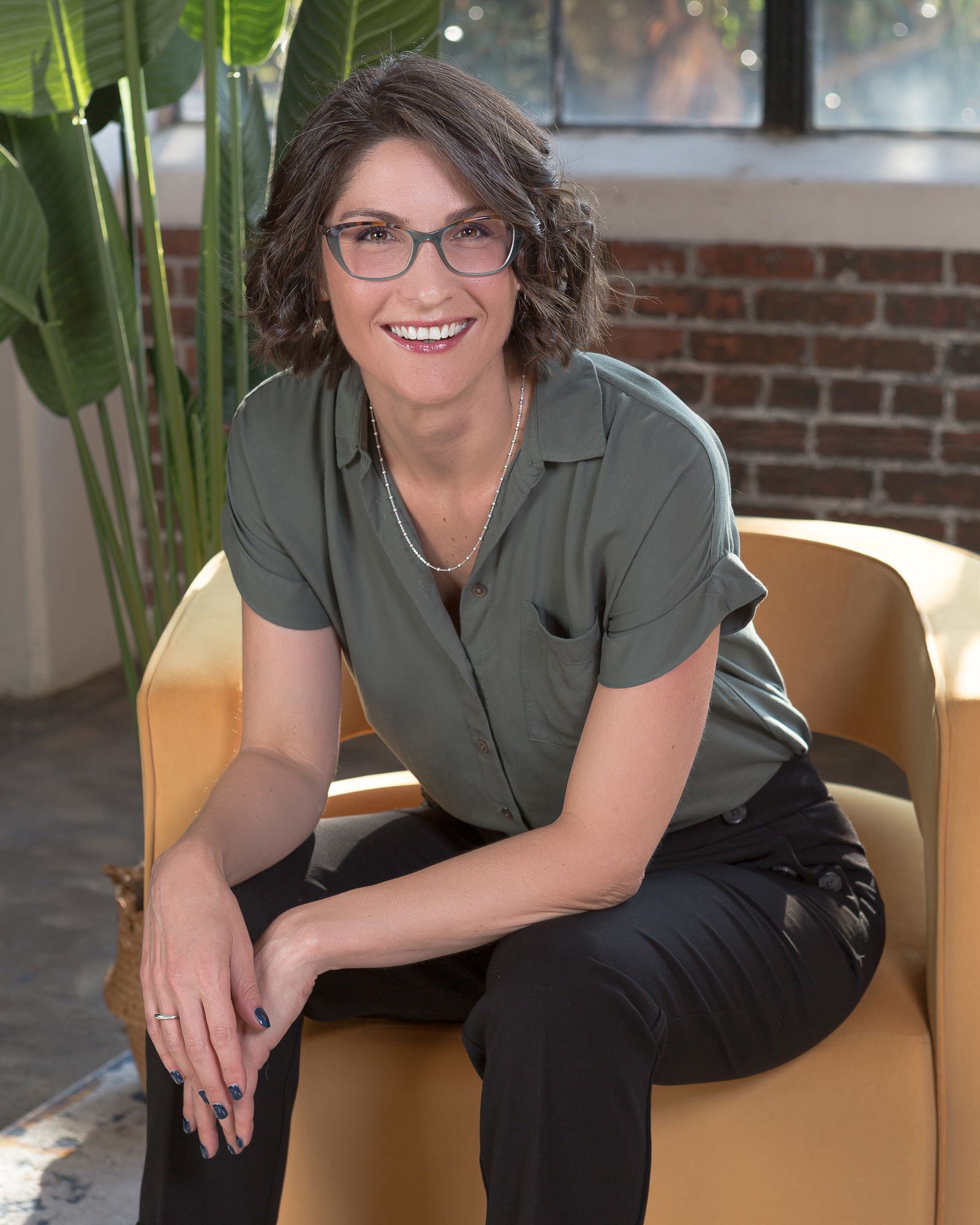 Headshot of Tory Gray with short brown hair and glasses smiling, seated on a yellow chair, wearing a green shirt and black pants, with plants nearby.
