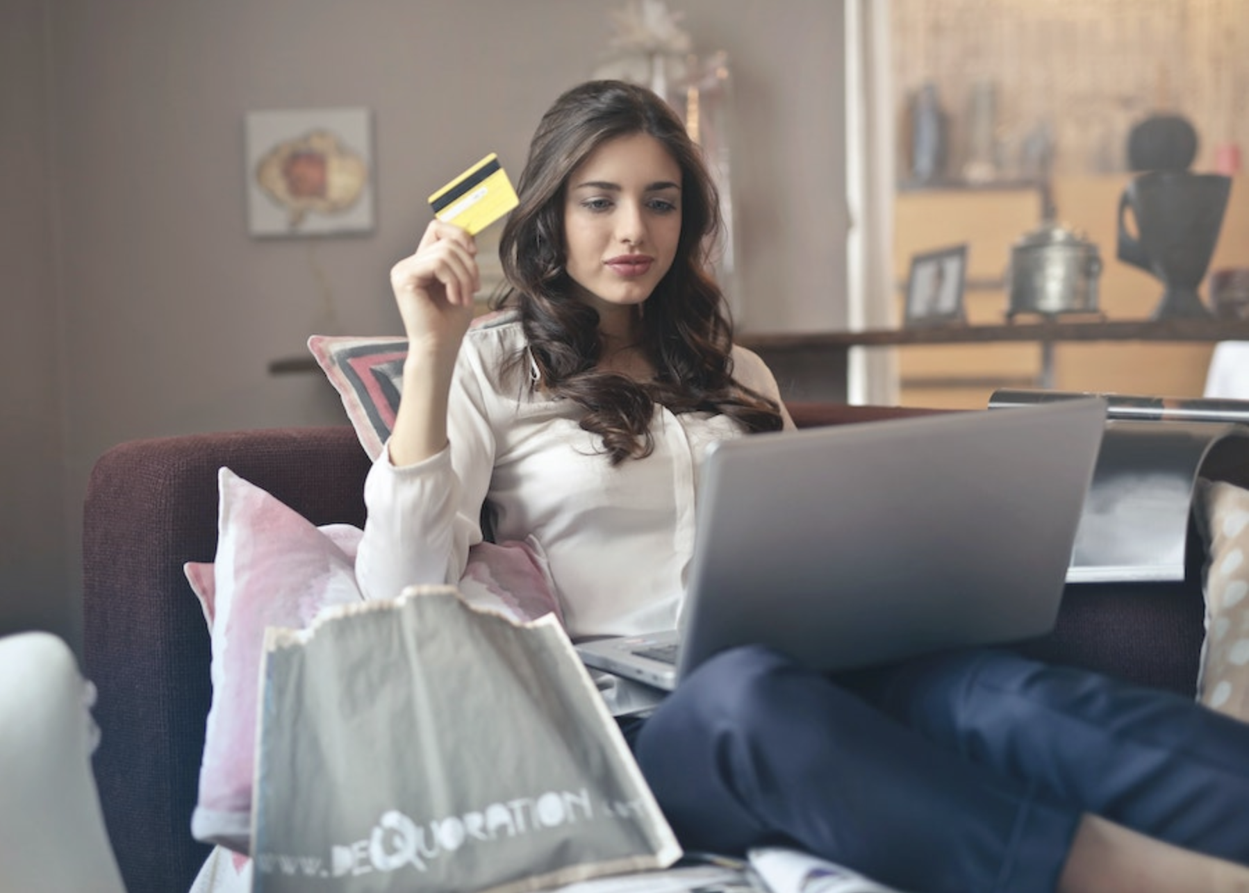 image of a brown-haired white woman sitting on a couch in front of a laptop, holding a credit card, about to buy something online from an ecommerce website
